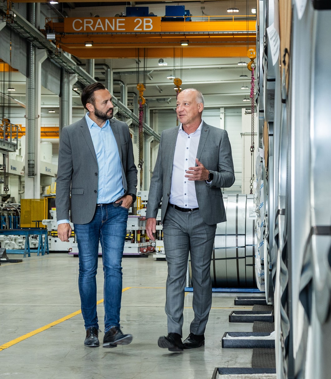 Zwei Führungskräfte im Gespräch in Produktionshalle mit Stahlcoils im Hintergrund.