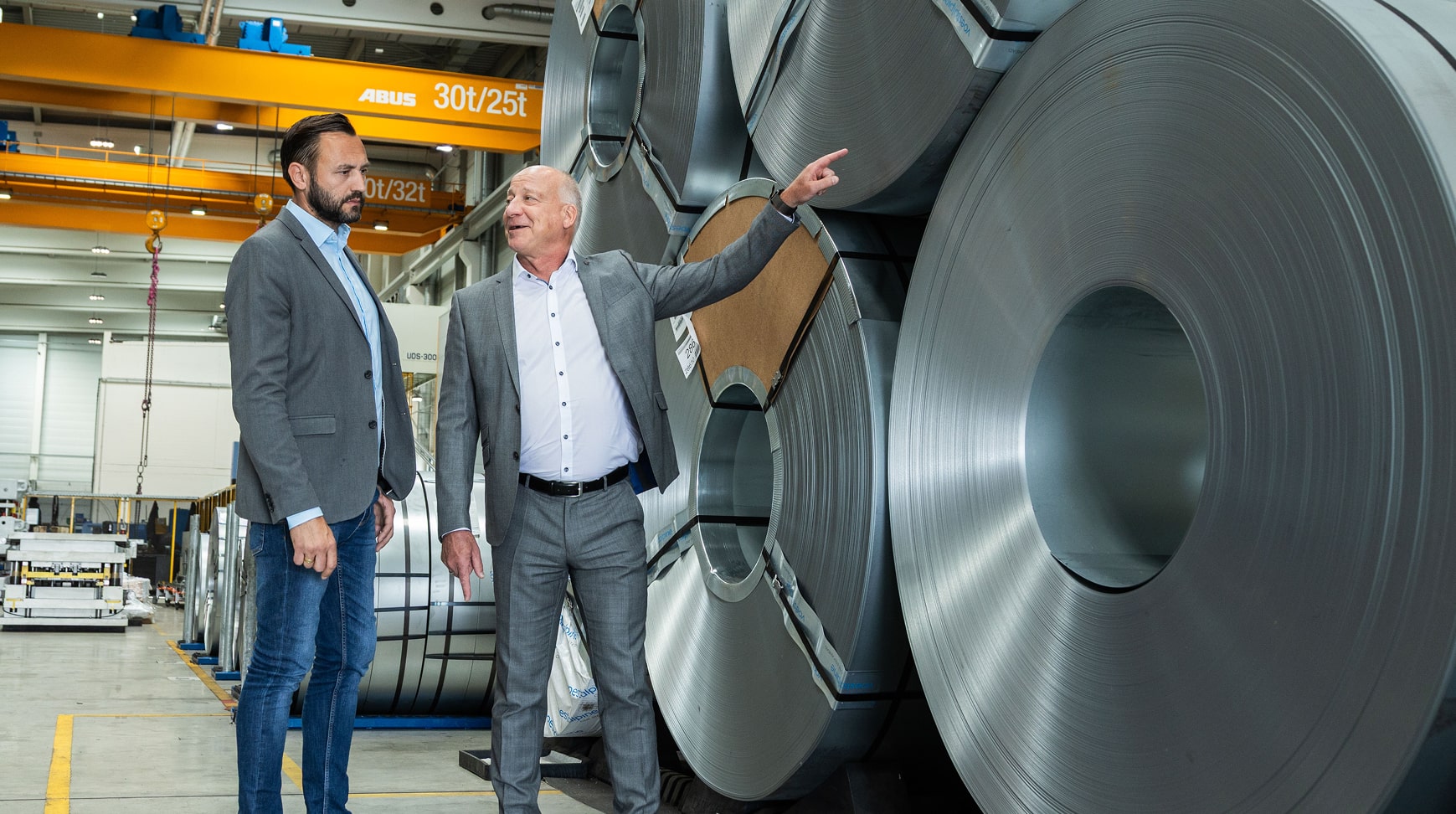 Zwei Geschäftsführer im Gespräch vor gestapelten Stahlcoils in einer Industriehalle.